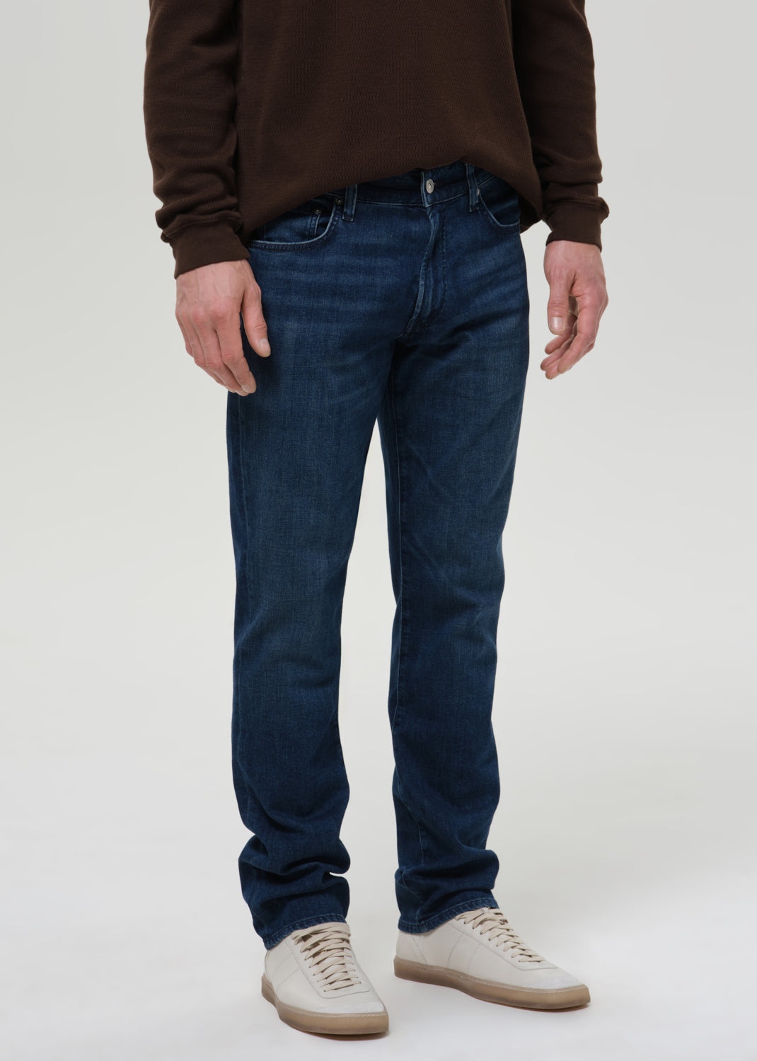 A person wearing dark blue slim-fit jeans, a brown long-sleeve top, and off-white sneakers, shown from the chest down against a grey background.