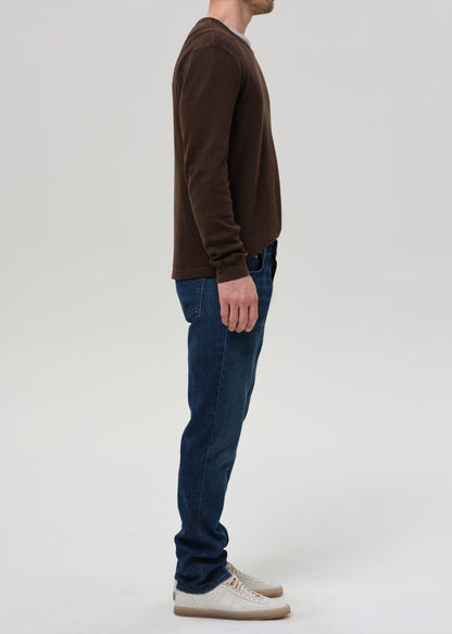 A man shown in profile wearing a dark brown long-sleeve crewneck sweater, dark blue jeans, and off-white sneakers, standing against a grey background.
