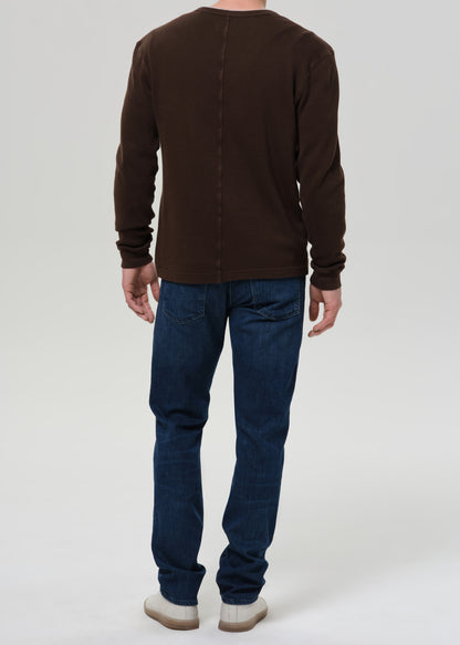 A man viewed from the back wearing a dark brown long-sleeve henley shirt and dark blue jeans, standing against a grey background.