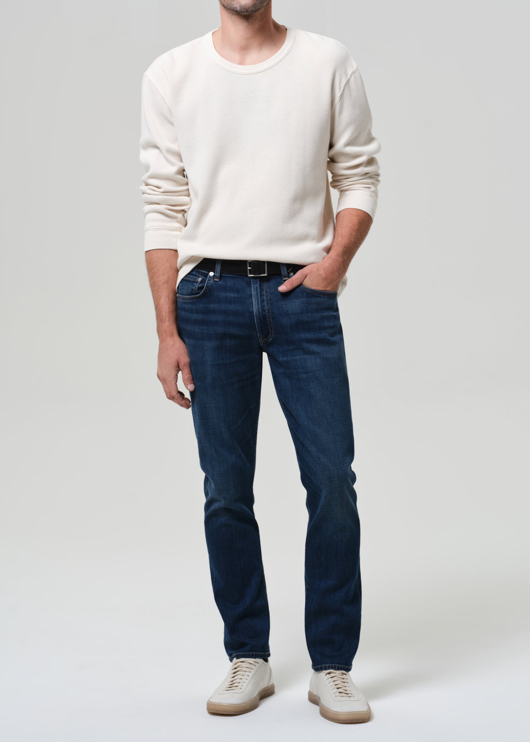 Man wearing a white sweater and dark blue jeans on a plain background
