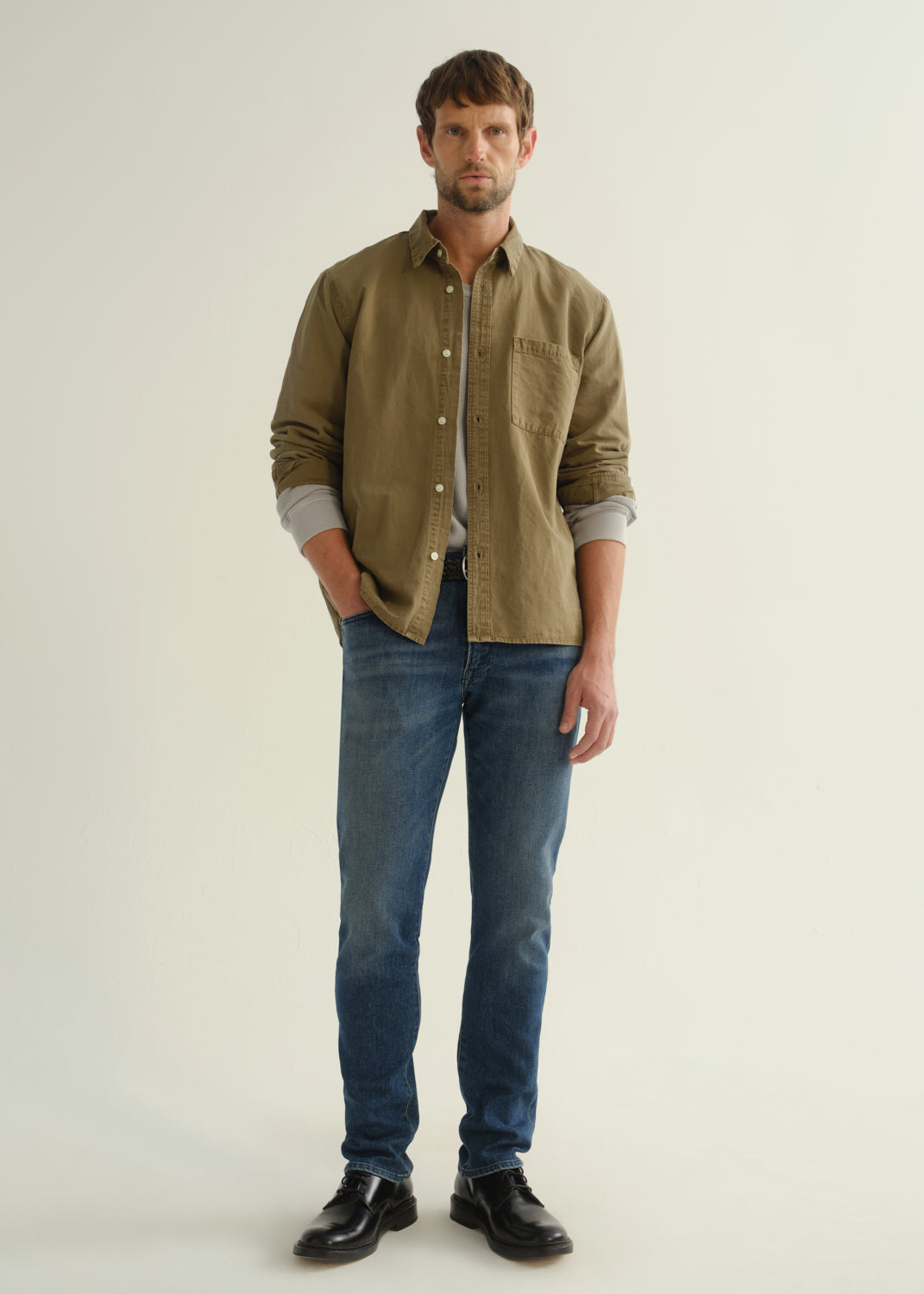 Person wearing an olive green button-front shirt, medium indigo jeans, and black boots, front full body view on a light grey background.
