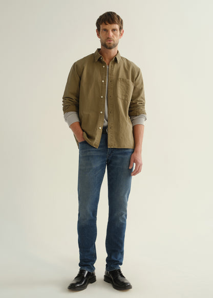 Person wearing an olive green button-front shirt, medium indigo jeans, and black boots, front full body view on a light grey background.
