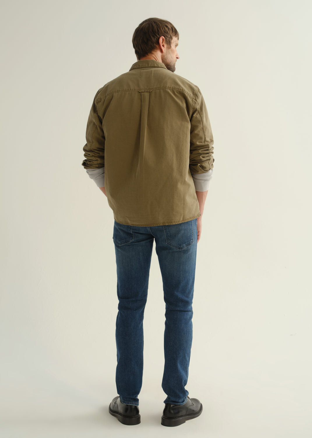 Person wearing an olive green button-front shirt, medium indigo jeans, and black boots, back full body view on a light grey background.