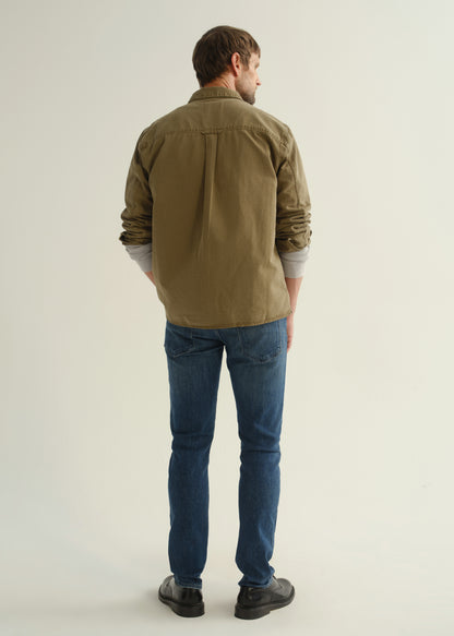 Person wearing an olive green button-front shirt, medium indigo jeans, and black boots, back full body view on a light grey background.