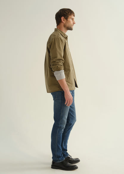 Person wearing an olive green button-front shirt, medium indigo jeans, and black boots, side full body view on a light grey background.