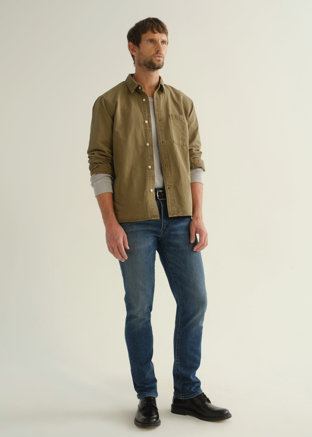 Person wearing an olive green button-front shirt, medium indigo jeans, and black boots, angled front full body view on a light grey background.