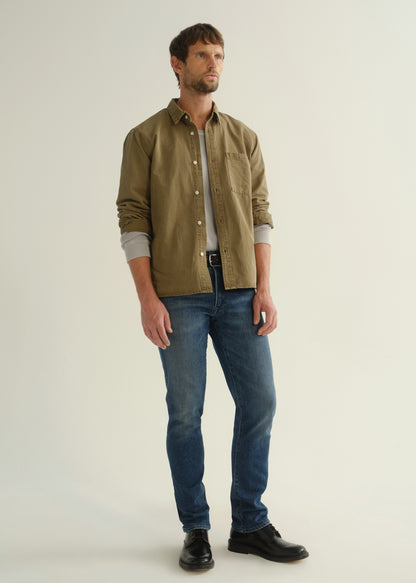Person wearing an olive green button-front shirt, medium indigo jeans, and black boots, angled front full body view on a light grey background.