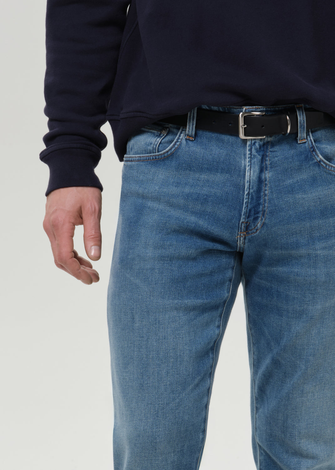 Close-up of a person wearing medium-wash blue jeans with a black belt and a dark navy sweatshirt, shown from the chest down against a grey background.