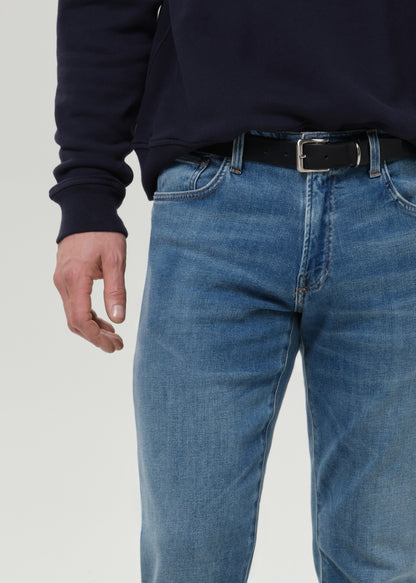 Close-up of a person wearing medium-wash blue jeans with a black belt and a dark navy sweatshirt, shown from the chest down against a grey background.