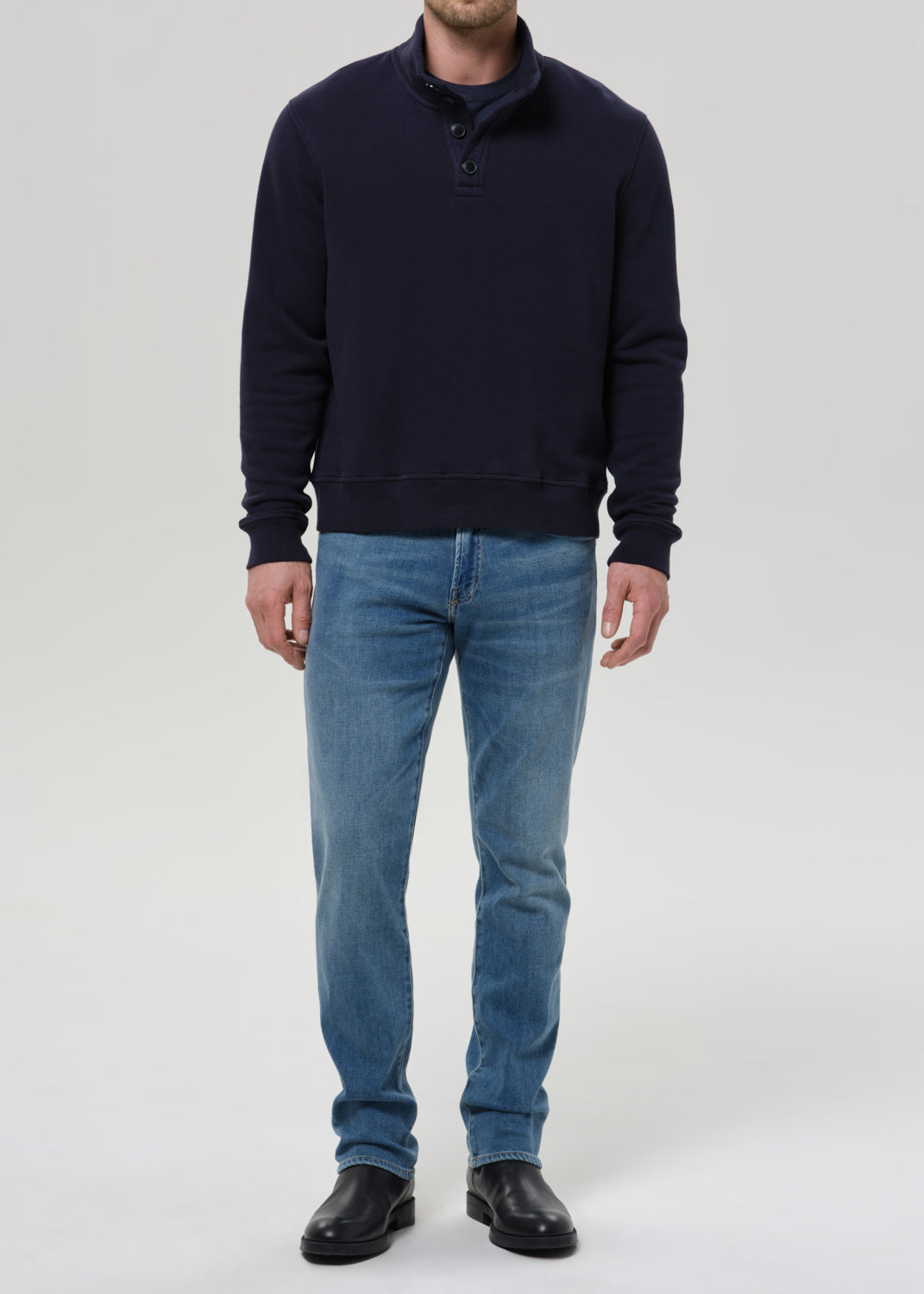 A man wearing a dark navy blue long-sleeve quarter-placket sweater, blue jeans, and black leather shoes, standing against a grey background.