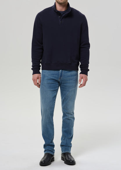 A man wearing a dark navy blue long-sleeve quarter-placket sweater, blue jeans, and black leather shoes, standing against a grey background.