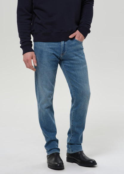A person wearing medium-wash blue jeans, a dark navy sweatshirt, and black leather boots, standing against a grey background.