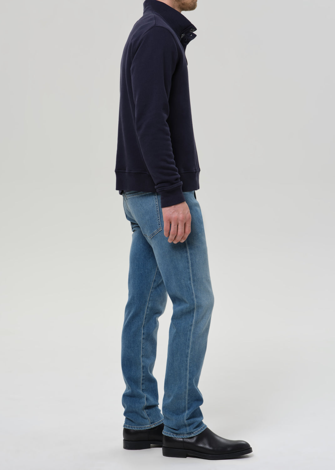 A man shown in profile wearing a dark navy quarter-zip fleece, light blue jeans, and black leather boots, standing against a grey background.