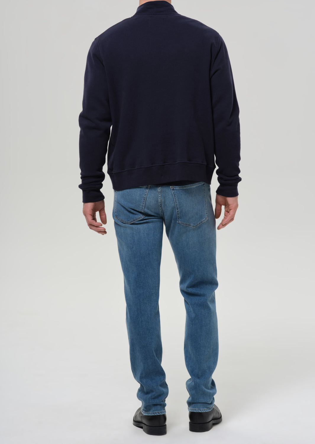 A man viewed from the back wearing a dark navy blue crewneck sweatshirt, blue jeans, and black shoes, standing against a grey background.