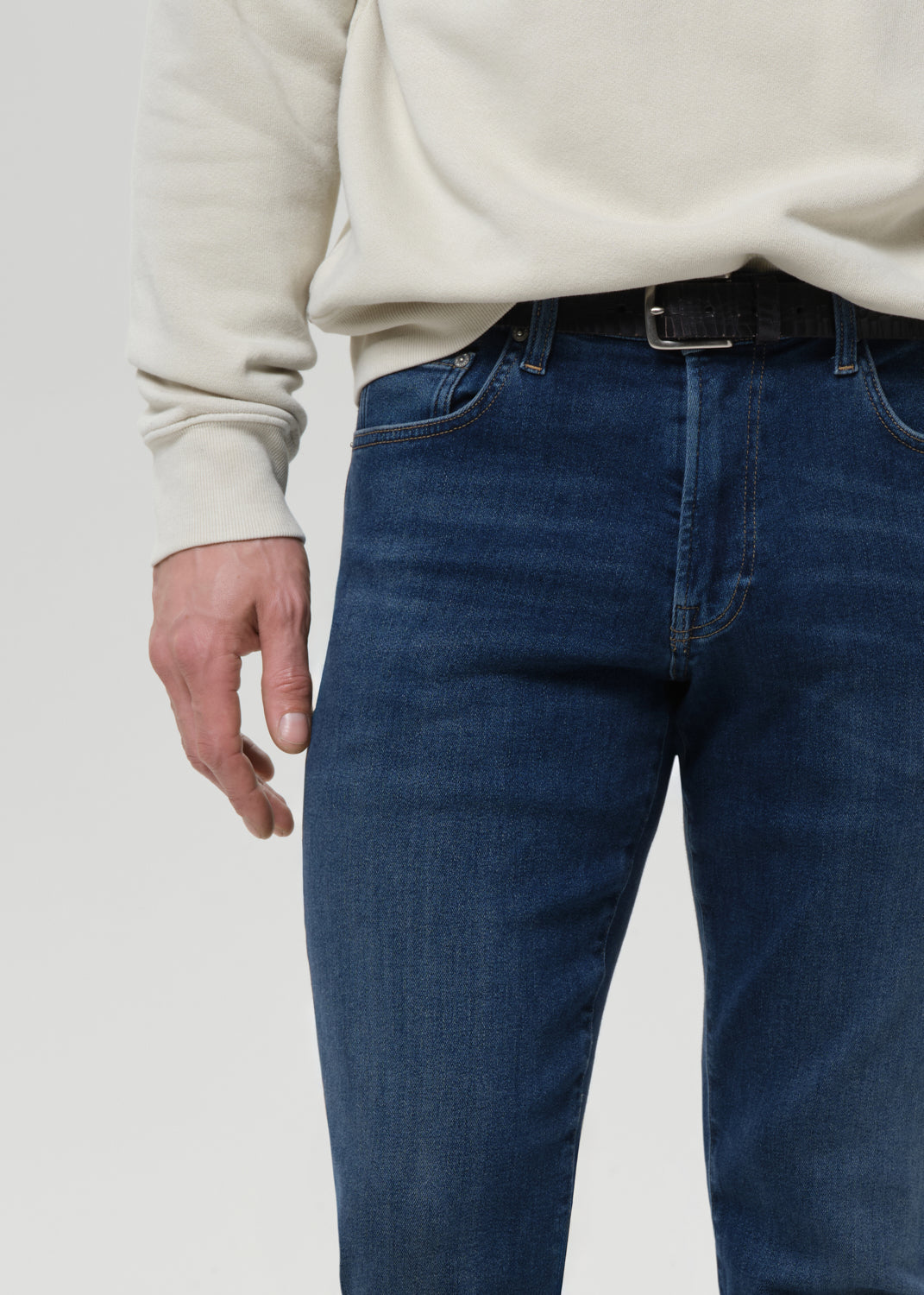 A close-up of a person wearing dark blue denim jeans with a black belt and a cream-colored sweatshirt, shown from the chest down against a grey background.