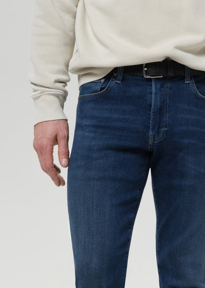 A close-up of a person wearing dark blue denim jeans with a black belt and a cream-colored sweatshirt, shown from the chest down against a grey background.