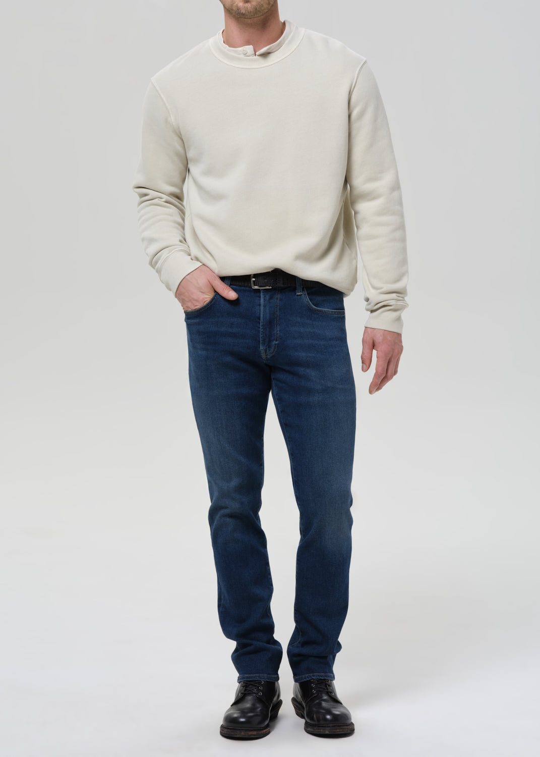 A man wearing a cream-colored long-sleeve crewneck sweater, dark blue jeans, and black leather shoes, standing against a grey background.