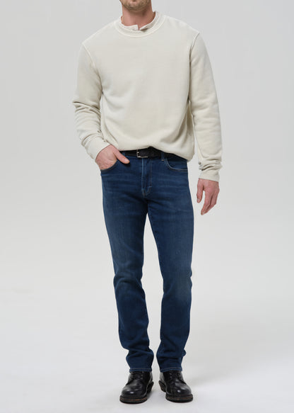 A man wearing a cream-colored long-sleeve crewneck sweater, dark blue jeans, and black leather shoes, standing against a grey background.