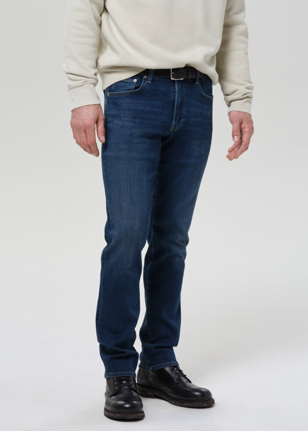 A person wearing dark blue slim-fit jeans, a cream sweatshirt, and black leather shoes, standing against a grey background.