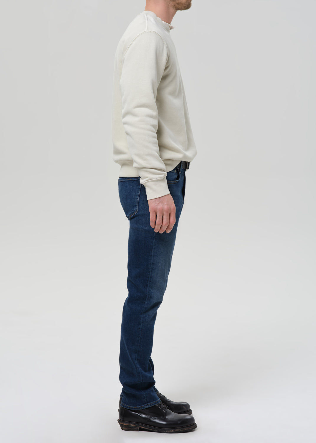 A man shown in profile wearing a cream long-sleeve knit sweater, dark blue jeans, and black leather shoes, standing against a grey background.