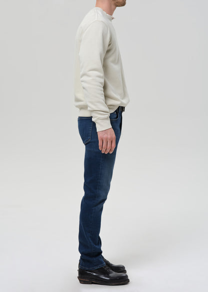 A man shown in profile wearing a cream long-sleeve knit sweater, dark blue jeans, and black leather shoes, standing against a grey background.