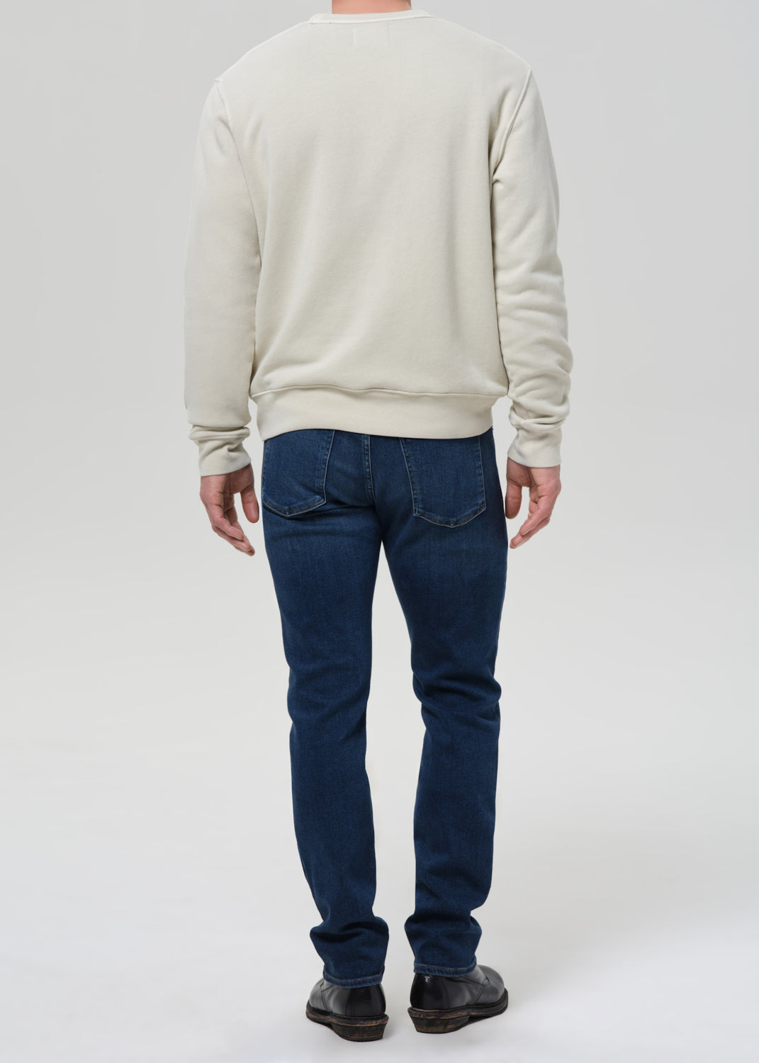 A man viewed from behind wearing a cream-colored crewneck sweatshirt, dark blue jeans, and black shoes, standing against a grey background.