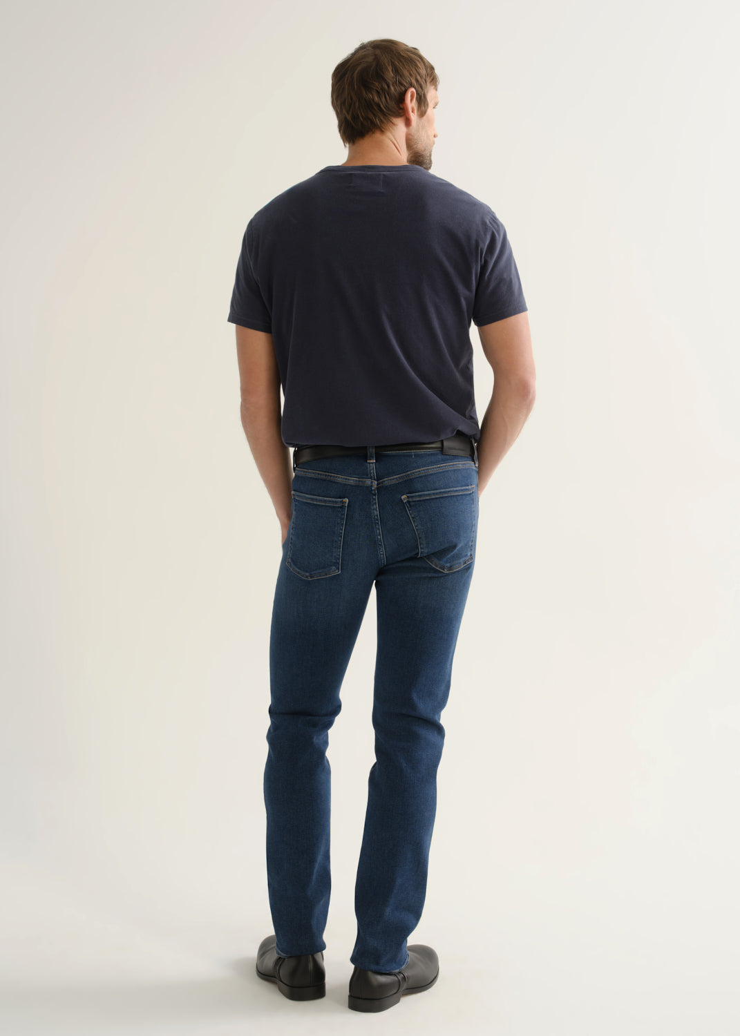Person wearing a dark navy short sleeve t-shirt, medium indigo jeans, and black boots, back full body view on a light grey background.