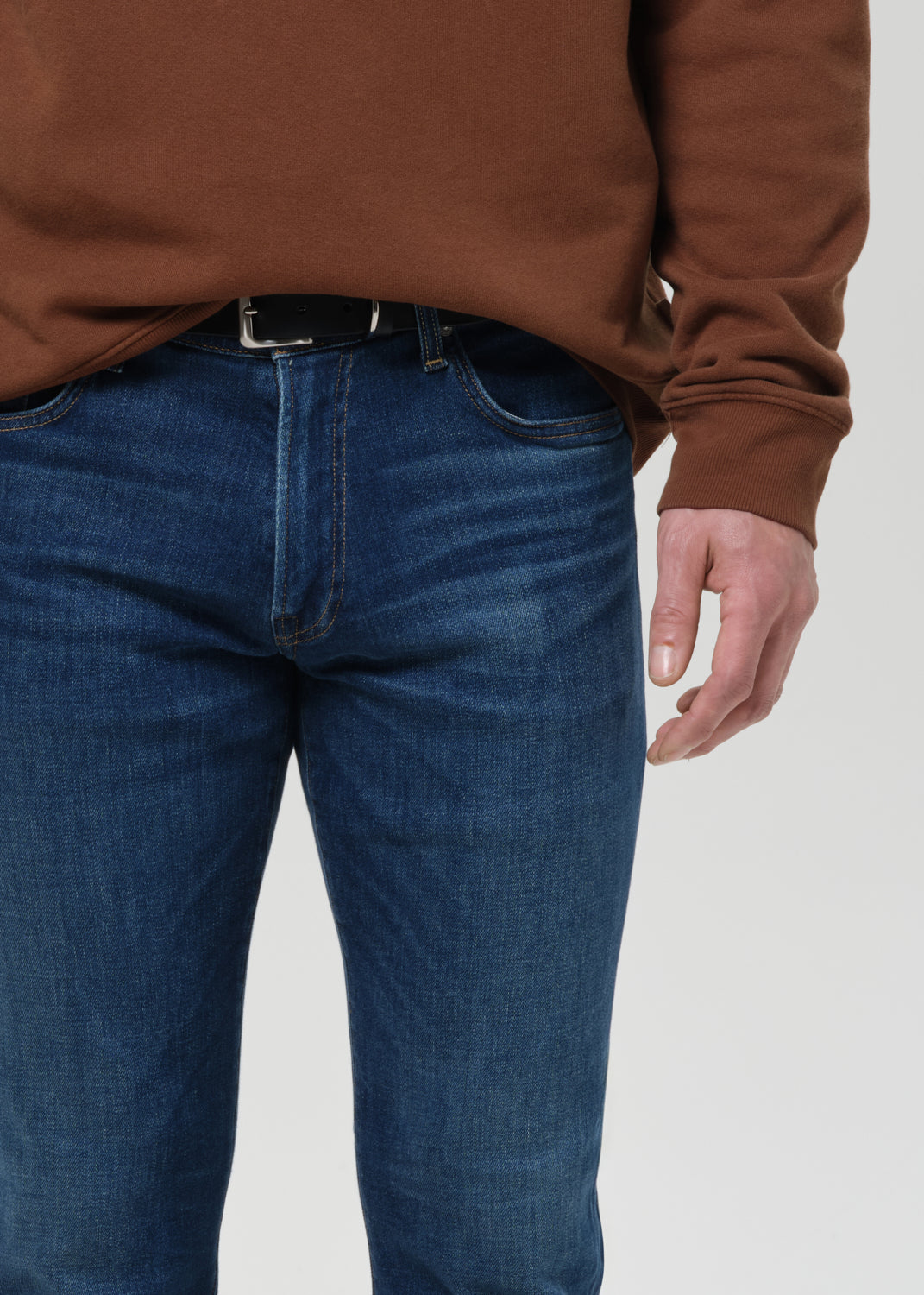 Close-up of a person wearing medium blue denim jeans with a black belt and a brown long-sleeve top, cropped from the chest down against a grey background.