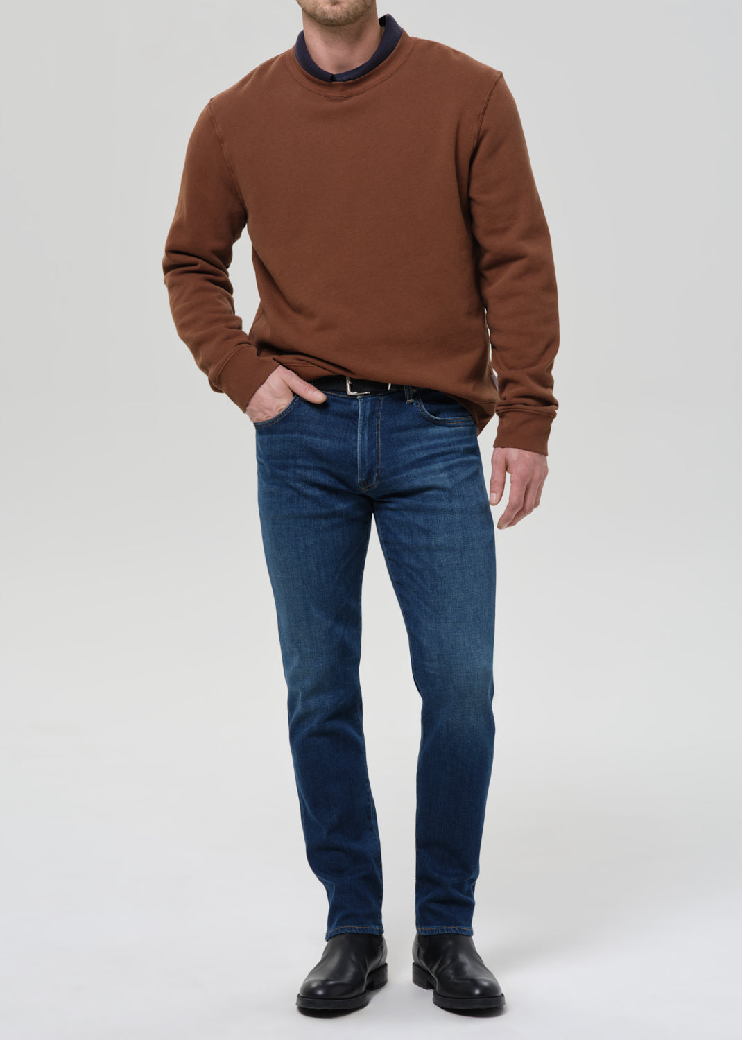 A man wearing a brown crewneck sweater with a dark navy collar, blue jeans, and black leather shoes, standing against a grey background.
