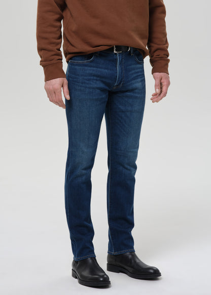 A person wearing dark blue slim-fit jeans, a brown sweatshirt, and black leather boots, standing against a grey background.