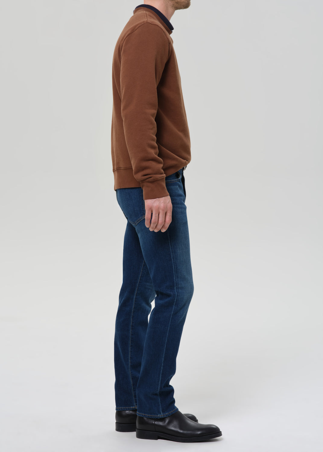 A man shown in profile wearing a brown long-sleeve crewneck sweater, blue jeans, and black leather shoes against a grey background.