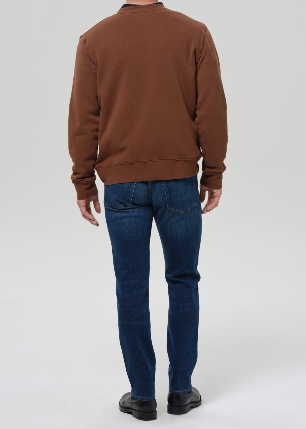 A man viewed from behind wearing a brown crewneck sweatshirt, dark blue jeans, and black shoes, standing against a grey background.