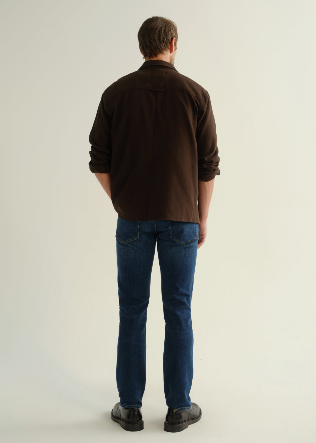 Person wearing a dark brown button-front shirt, medium indigo jeans, and black boots, back full body view on a light grey background.