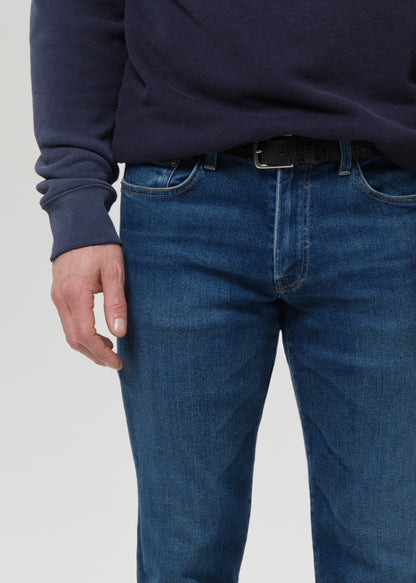 A close-up of a person wearing blue denim jeans with a dark belt and a navy long-sleeve top, cropped from the chest down against a grey background.