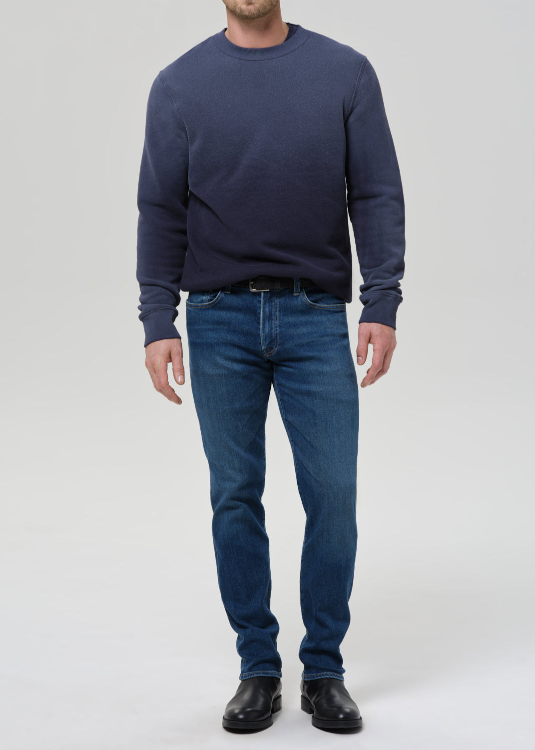 A man wearing a navy blue crewneck sweatshirt, dark blue jeans, and black leather shoes, standing against a grey background.