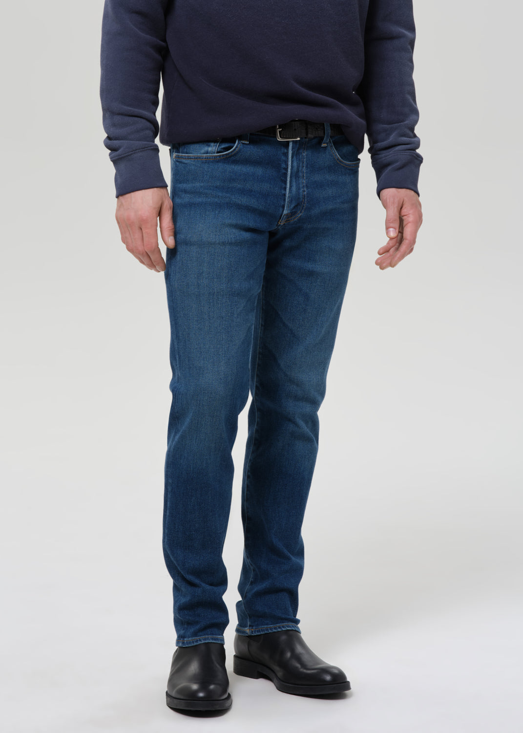 A man wearing a navy blue long-sleeve shirt, blue slim-fit jeans, and black leather boots, standing against a grey background.