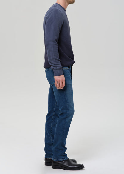 A man shown in profile wearing a dark navy long-sleeve crewneck sweater, blue jeans, and black leather shoes, standing against a grey background.
