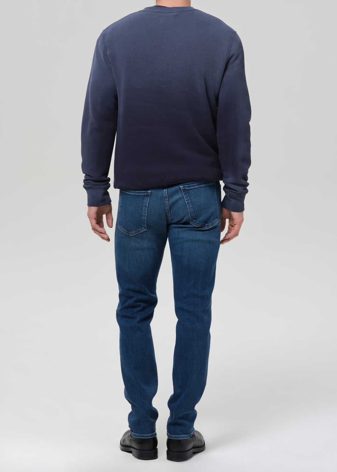 A man viewed from the back wearing a dark navy blue long-sleeve crewneck sweatshirt and blue jeans, standing against a grey background.