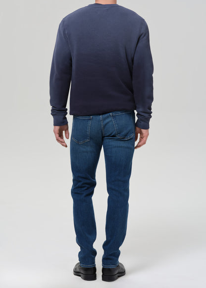 A man viewed from the back wearing a dark navy blue long-sleeve crewneck sweatshirt and blue jeans, standing against a grey background.