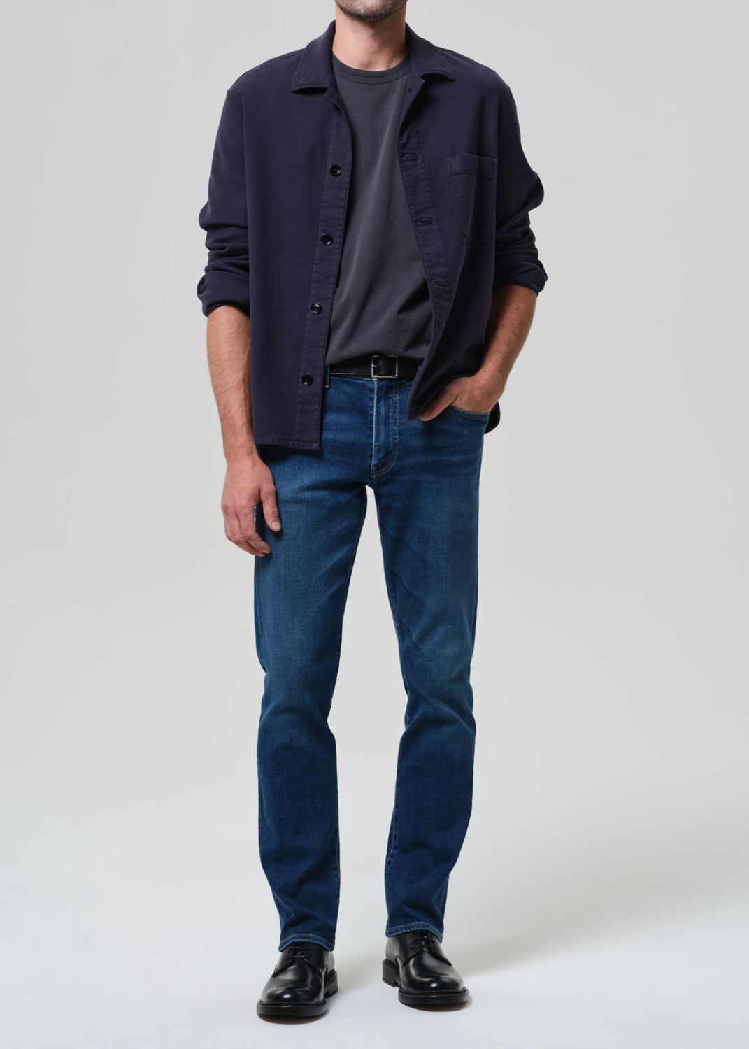 Man wearing a navy shirt, dark gray t-shirt, blue jeans, and black shoes on a grey background