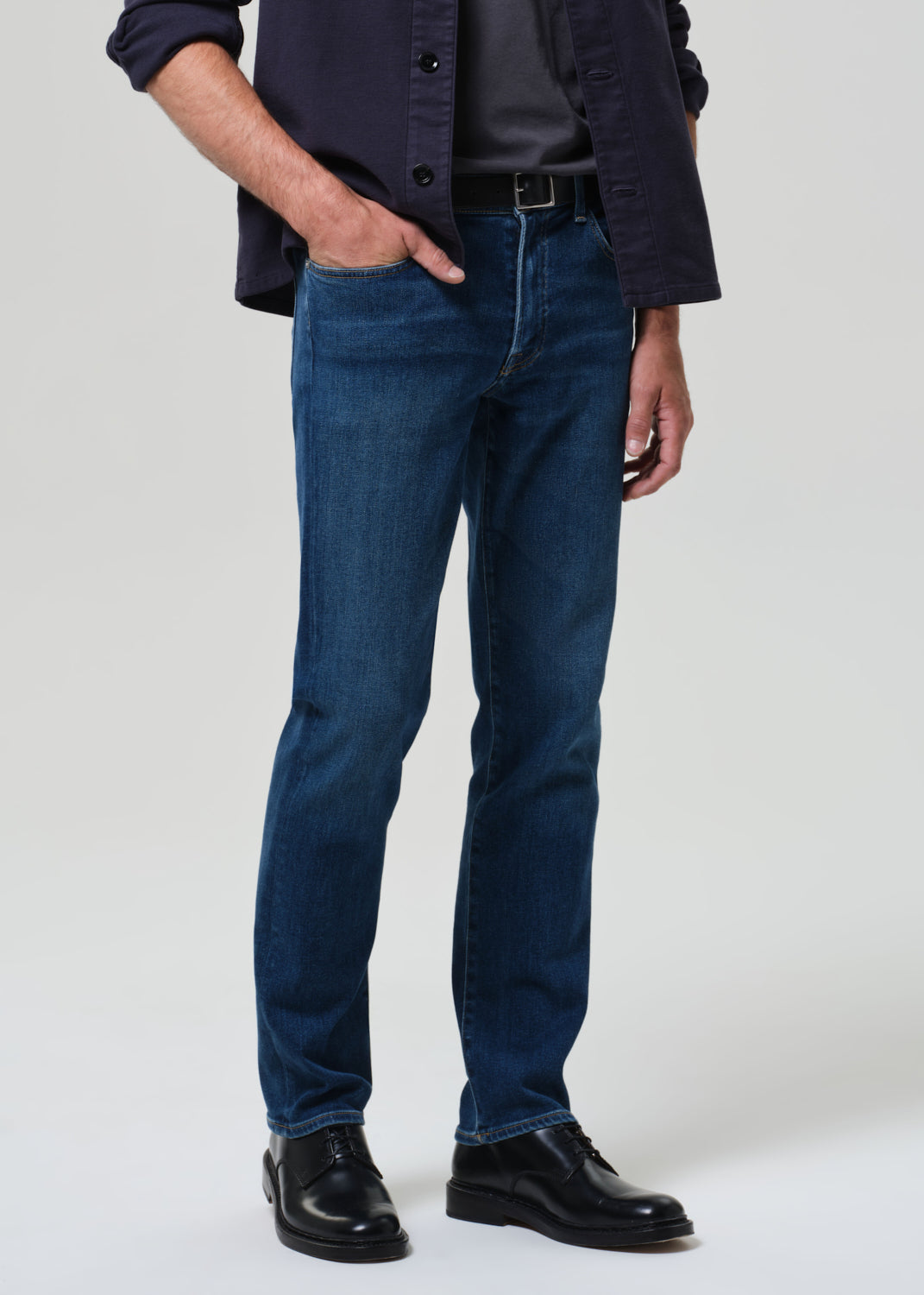 Man wearing a navy shirt, dark gray t-shirt, blue jeans, and black shoes on a grey background