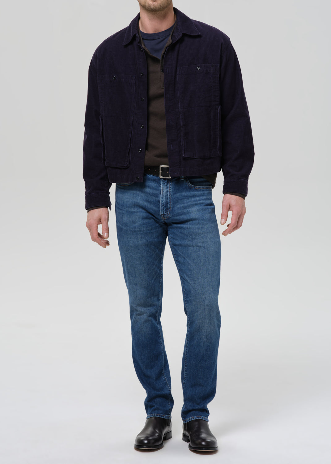 A man wearing a dark navy corduroy jacket over a dark brown shirt, paired with blue jeans and black leather boots, standing against a grey background.
