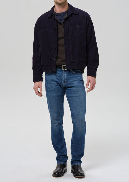 A man wearing a dark navy corduroy jacket over a dark brown shirt, paired with blue jeans and black leather boots, standing against a grey background.