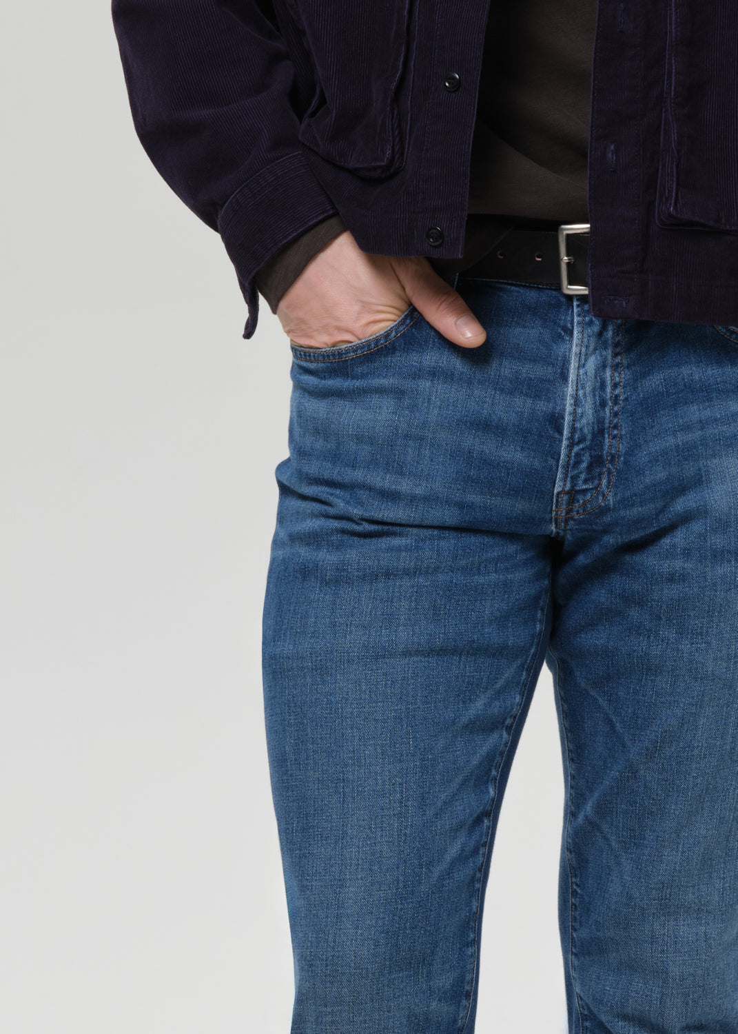 Close-up of a person wearing blue denim jeans with a black belt, dark brown shirt, and a dark navy corduroy jacket, standing against a grey background.