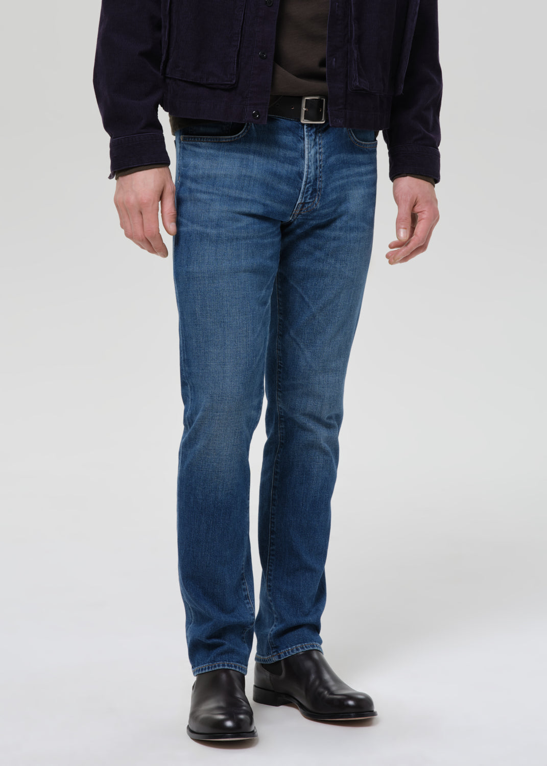 A person wearing blue denim jeans, a dark navy jacket, a dark shirt, and black leather boots, standing against a grey background.