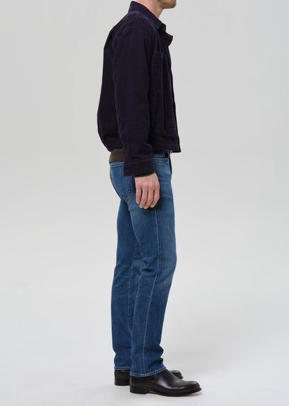 A man shown in profile wearing a dark navy corduroy jacket, blue denim jeans, and black leather shoes, standing against a grey background.