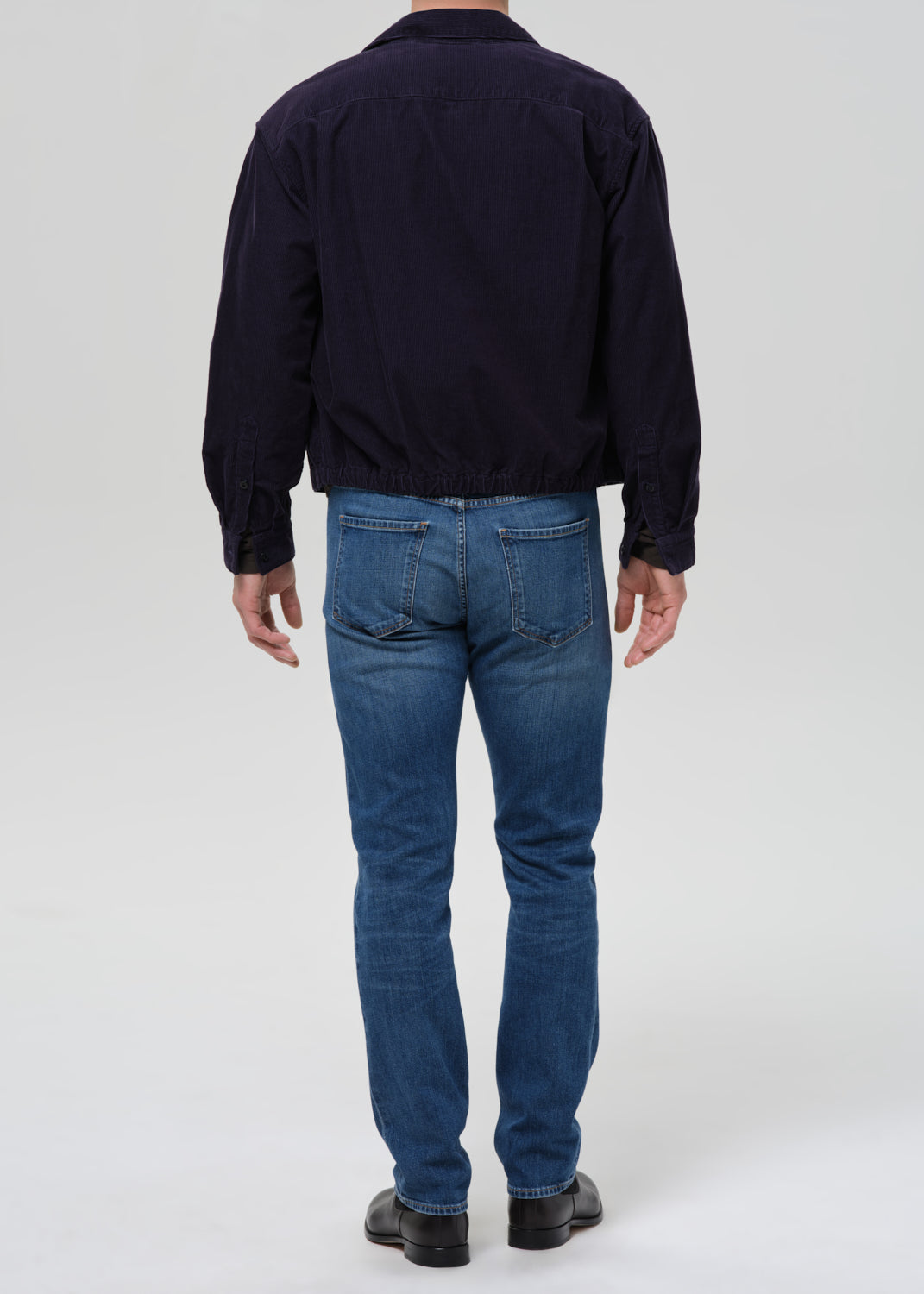 A man viewed from behind wearing a dark navy jacket, blue jeans, and black shoes, standing against a grey background.