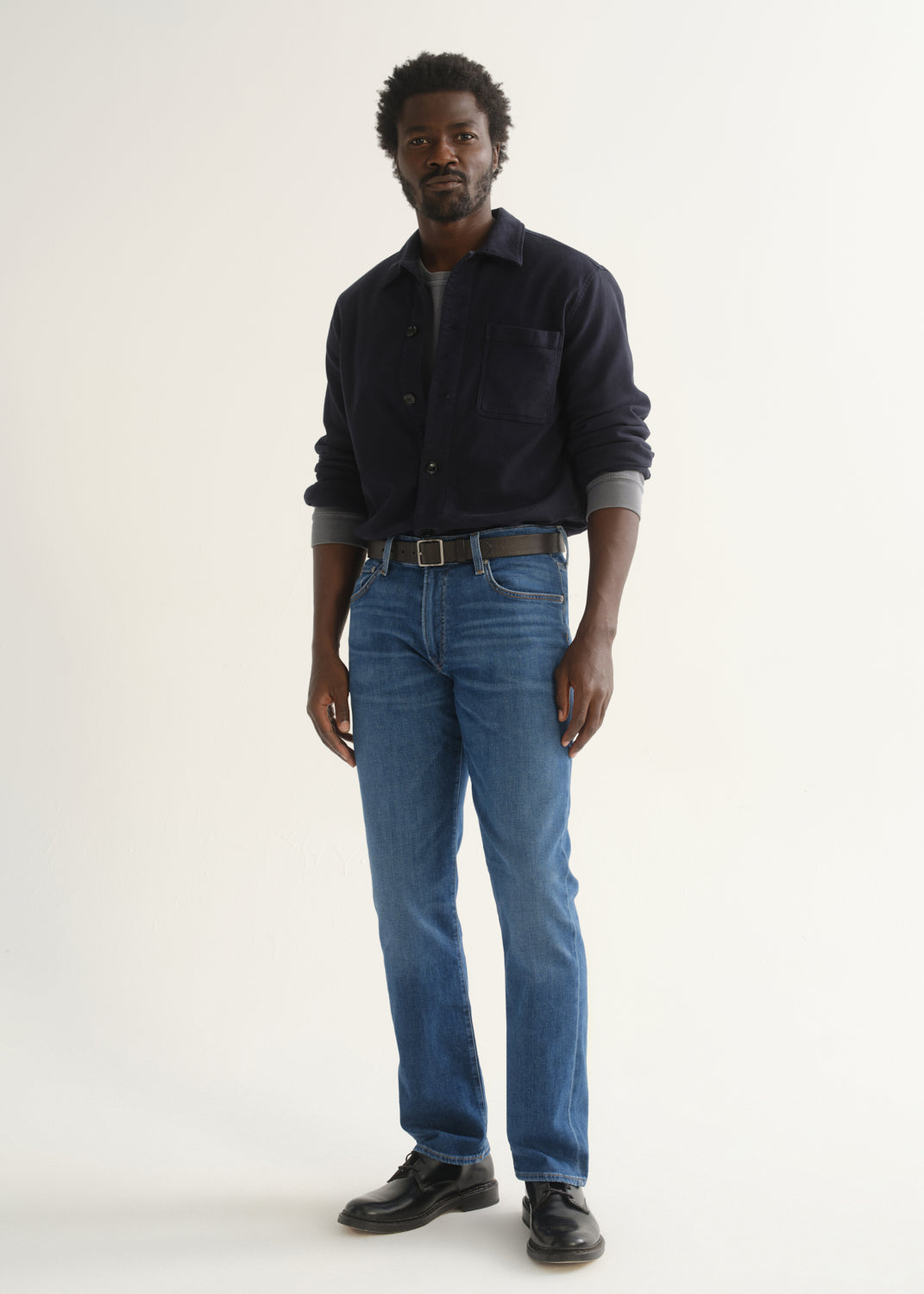 Person wearing medium blue straight leg jeans with a navy long sleeve collared pullover and black shoes, front full body view on a light grey background.