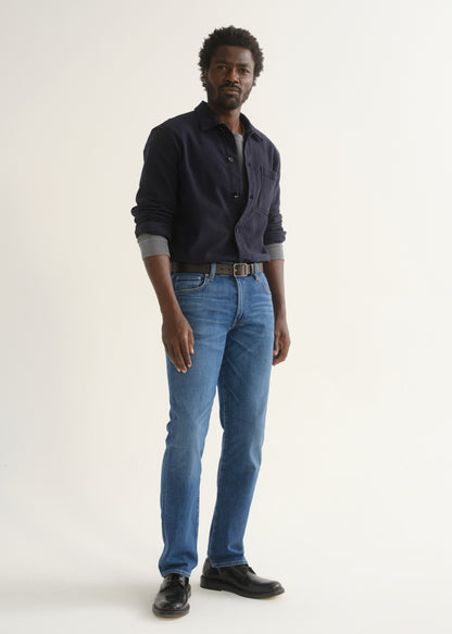 Person wearing medium blue straight leg jeans with a navy long sleeve collared pullover and black shoes, angled front full body view on a light grey background.
