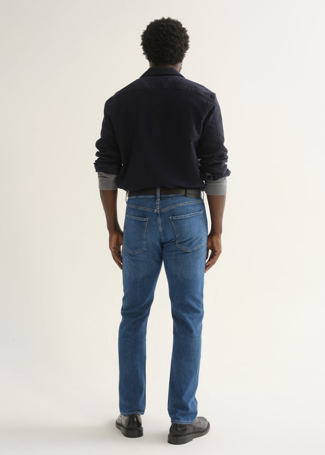 Person wearing medium blue straight leg jeans with a navy long sleeve collared pullover and black shoes, back full body view on a light grey background.